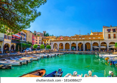 Puerto antiguo de Porto Vecchio con barcos a motor sobre agua turquesa, árboles verdes y edificios tradicionales en el centro histórico de la ciudad de Desenzano del Garda, fondo del cielo azul, Lombardía, norte de Italia