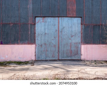 Old High Groove Metal Gate With Peeling Grey Paint Of An Old Warehouse With Old Metal-covered Facade On A Cold Sunny Day.