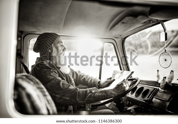 Old hipster man in a\
van  looking at a road map, he is tattooed and he wears a white\
beard. black and white