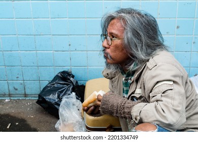 An Old, Homeless, Long-bearded Asian Man Sits On The Street Eating Bread After Receiving A Delicious Snack. From People Passing By The Poor Man Had No Home In The Streets.