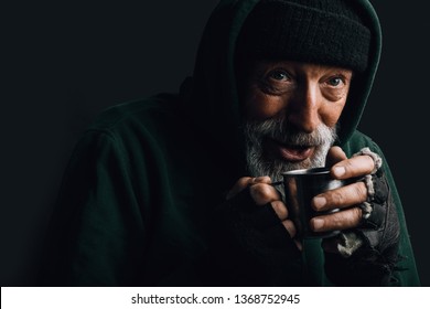 Old homeless man with grey beard covering up in green decrepit wear holding a mug of hot tea to warm himself in a cold night