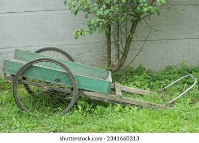 old homemade retro wooden green color manual with bicycle wheels easy to transport goods wheelbarrow stands on green grass on the street in the afternoon near a gray fence and a tree in summer