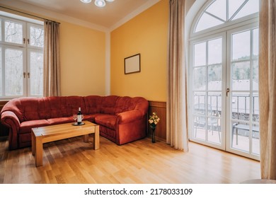 Old hotel living room with red couch and wooden coffee table. In room can be found balcony window in arc shape, wooden floor and yellow-painted walls. There are also little additions as wine set.