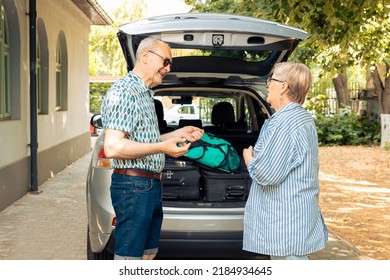 Old Husband And Wife Sitting In Driveway With Car, Leaving On Holiday Vacation Adventure After Loading Travelling Bags And Suitcases In Automobile Trunk. Leaving On Journey With Trolley For Leisure.