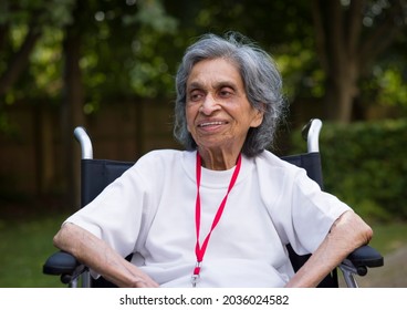 Old Indian Woman Sitting In A Wheelchair, Smiling. Outdoors In A Park Or Garden In Summer UK