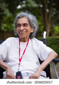 Old Indian Woman Sitting In A Wheelchair, Smiling. Outdoors In A Park Or Garden In Summer UK