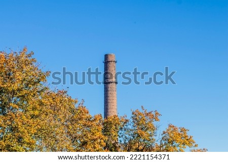 An old industrial brick chimney of a factory behind yellow autumn trees with clear blue sky and copy space in the background