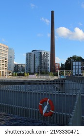 An Old Industrial Smokestack And Some Modern Buildings. A Red And White Lifesaver In The Foreground. Ranta-Tampella, Tampere, Finland. August 12th 2022.