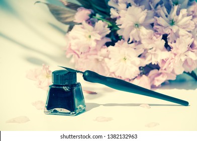 Old ink pen and ink bottle on white background. Vintage calligraphy pen and bottle of ink. Selective focus. Tender pink flowers in the background. Cherry blossom twig with black ink