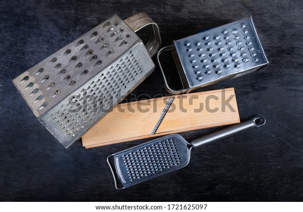 Old kitchen box graters with\
multiple grating surfaces and different holes, flat grater and\
wooden grater with special stainless steel blade on the black\
surface\
