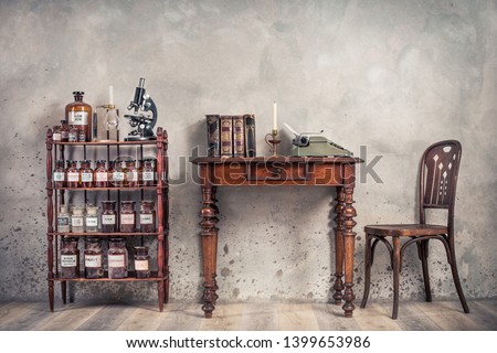 Old laboratory bottles, vintage microscope, glassware support on the shelving, classic typewriter and antique books on wooden table and chair front concrete wall background. Retro style filtered photo