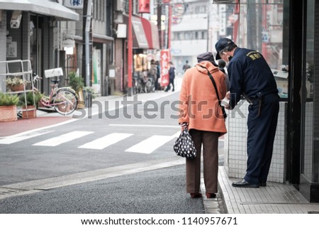 An old lady is asking direction and assistance from a local police on the street of japan. The policeman is wearing his uniform showing 'Policeman' on the back.