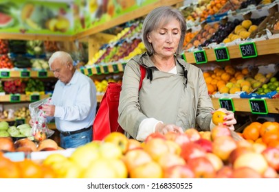 Old Lady Choosing Mandarins While Standing In Salesroom Of Greengrocer. Old Man Shopping In Background.