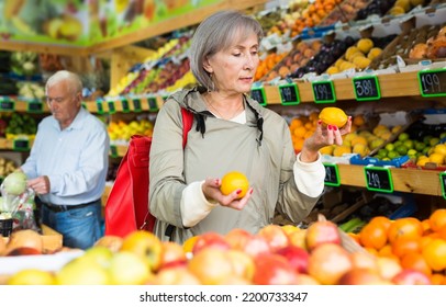 Old Lady Choosing Mandarins While Standing In Salesroom Of Greengrocer. Old Man Shopping In Background.