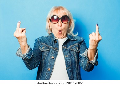 An old lady in a denim jacket, sunglasses makes an unpretentious gesture with the middle finger on a blue background. Concept fashionable grandma, old woman.
