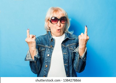 An Old Lady In A Denim Jacket, Sunglasses Makes An Unpretentious Gesture With The Middle Finger On A Blue Background. Concept Fashionable Grandma, Old Woman.