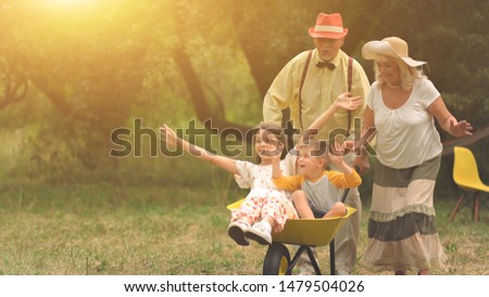The old lady and gentleman are pushing their grandchildren in a wheelbarrow.Funny boy and girl raised their hands up, their granparents are looking at them. The portrait of the ideal hapiness.