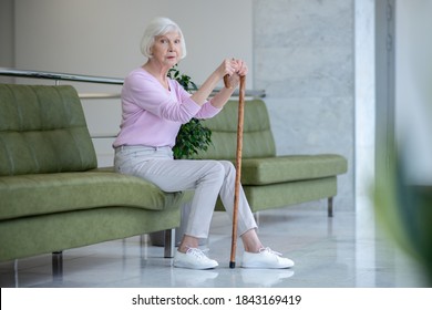 Old Lady. Grey-haired Woman With A Walking Stick Sitting In The Corridor And Looking Serious