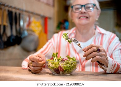 Old Lady Having Healthy Snack And Glass Of Water. Senior Woman Eating Fresh Salad At Her Wooden Kitchen Table. Copy Space