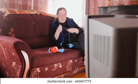 Old lady pensioner at home in glasses knitting in front of the TV