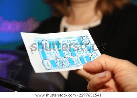 	
Old lady playing Bingo. Elderly woman's hands holding cardboard from bingo game at an event. Bingo player. Living room game. Classic board game. Entertainment. Woman's hands.