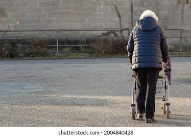 Old Lady, Senior Woman With Rollator For A Walk On The Street. Grandma With Wheeled Walker. Architecture Background Crumbling Due To Neglect In A Depressed Area District. Old-age Poverty Concept