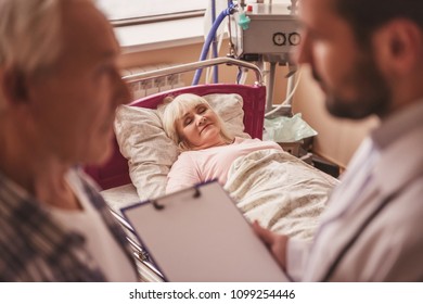 Old Lady Is Sleeping In Bed In The Hospital Ward While Her Husband And Doctor Are Talking