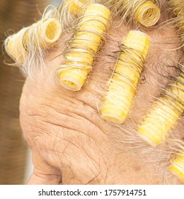 Old Lady With Sparse Thin Wispy Grey Hair Has Set Her Hair By Rolling It Up In Self Retaining Yellow Curlers. She Has Lines And Wrinkles On Her Forehead And Matt Makeup. Close Up View.