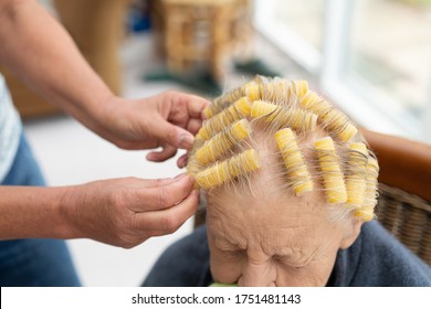 Old Lady With Thin Grey Hair Has Her Hair Set By A Family Member Using Self Retaining Yellow Curlers. She Sits On A Dining Chair In A Makeshift Home Salon During Covid-19 Lock Down. 