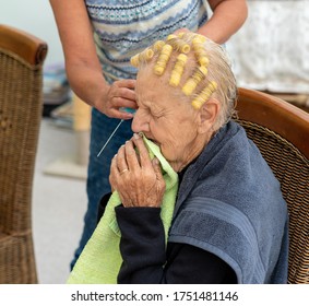 Old Lady With Thin Grey Hair Has Her Hair Set By A Family Member Using Self Retaining Yellow Curlers. She Sits On A Dining Chair In A Makeshift Home Salon During Covid-19 Lock Down. 