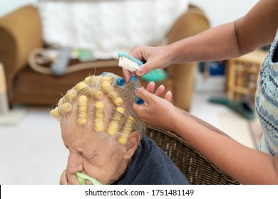 Old Lady With Thin Grey Hair Has Her Hair Set By A Family Member Using Self Retaining Yellow Curlers. She Sits On A Dining Chair In A Makeshift Home Salon During Covid-19 Lock Down. 