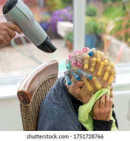 Old Lady With Thin Grey Hair Rolled In Self Retaining Yellow, Pink And Blue Curlers Covers Her Face With A Green Towel Under The Home Salon Hairdryer. Sits On A Dining Chair During Covid-19 Lockdown.