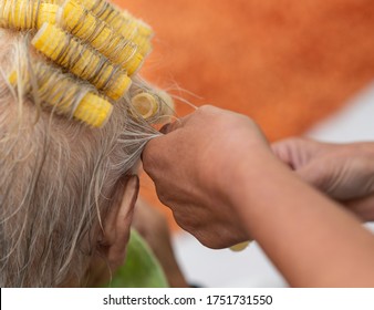 Old Lady With Thin Grey Hair Has Her Hair Set By A Family Member In A Makeshift Home Salon During The Covid-19 Lock Down. Close Up Of Self Retaining Yellow Curlers, Shallow Depth Of Field. 