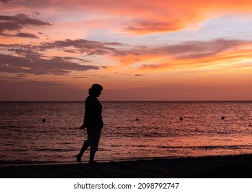 And Old Lady Walking On The Beach, With An Orange Sunset Behind Her