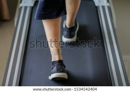 The old lady is wearing the sport shoes, doing the low impact exercise which is walking on the treadmill at home.