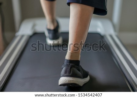 The old lady is wearing the sport shoes, doing the low impact exercise which is walking on the treadmill at home.