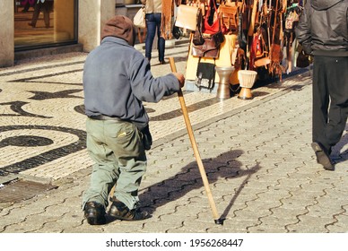 Old Lame Tramp Walks Holding A Stick.
Back View Of A Poor Man Trudging On A Shopping Street.