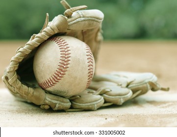 An Old Leather Baseball Mitt, Or Glove With A Worn Baseball Laying On A Home Plate. There Is Clay Around. Home Plate Needs To Be Dusted Off. Vintage Filter Applied. Shallow Depth Of Field.