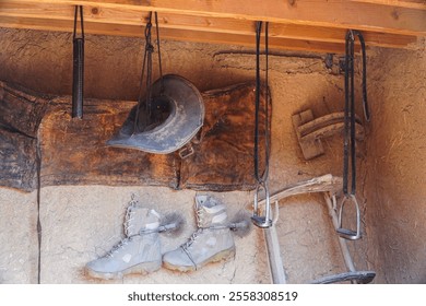 Old leather horse tack, boots, and a hat hang in a rustic stable. Rustic Western Horse Tack and Gear Hanging in Old Stable. - Powered by Shutterstock