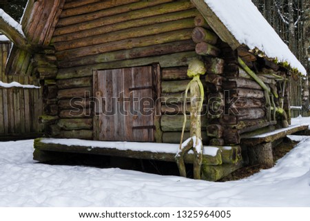 Old log house between trees in the winter. Vintage agriculture equipment hanging on the timber wall. Human shape sculpture.Mossy roof covered with snow. Tavern farm in Mustjoe, Estonia, Baltic, Europe