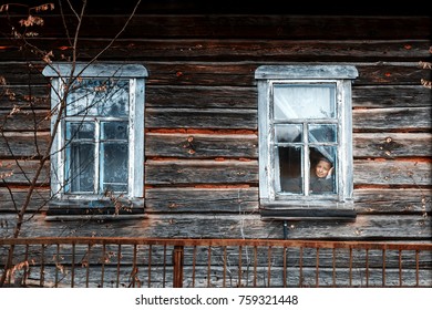 Old And Lonely Lady. Elderly Lonely Woman With Sadly Looking Through The Window. Very Old Wooden House.