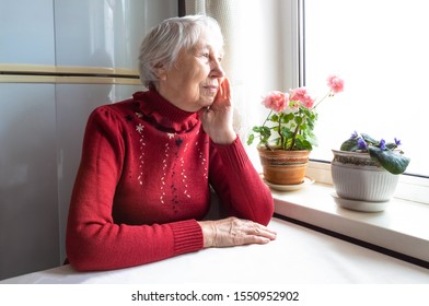 Old Lonely Woman Sitting Near The Window At Home. Concept Of Quarantine, Coronavirus Covid-19. Danger For Old People