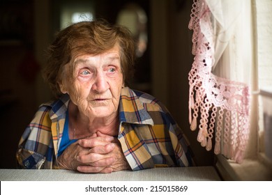 Old Lonely Woman Sitting At A Table Near The Window In His House. Old Age. Loneliness. 