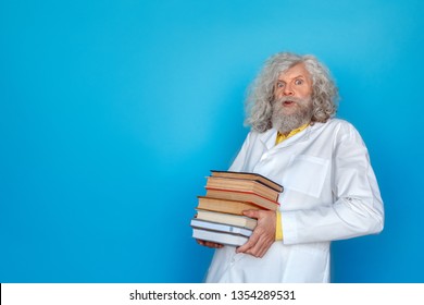 Old long-haired man wearing doctor's gown studio isolated on blue wall standing holding pile of books leaning back looking camera shocked