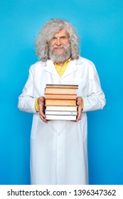 Old Long-haired Man Wearing Doctor's Gown Studio Isolated On Blue Wall Standing Holding Pile Of Books Full Body Shot Looking Camera Smiling Cheerful