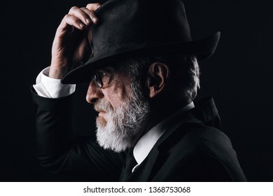 An Old Male Actor Playing An English Spy Role, Dressed In Black Suit, Hiding Behind Fedora Hat, Having Mysterious Elegant Look. Isolated Studio Portrait Over Dark Background