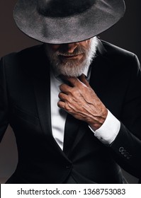 An Old Male Actor Playing An English Spy Role, Dressed In Black Suit, Hiding Behind Fedora Hat, Having Mysterious Elegant Look. Isolated Studio Portrait Over Dark Background