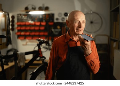 Old male bicycle mechanic doing his professional work in workshop using voice assistant on mobile phone, talking to smartphone. Copy space - Powered by Shutterstock