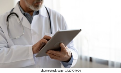 Old Male Doctor Holding Modern Digital Tablet Computer Standing In Hospital, Close Up View. Senior Middle Aged Professional Medic Wears White Coat And Stethoscope Using Healthcare Ehealth Application.