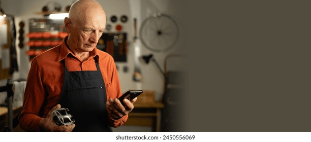 An old male repairman is repairing a bicycle in his garage, holding a pedal in his hands and looking for information about a breakdown on the Internet using a smartphone. Close-up. Banner - Powered by Shutterstock